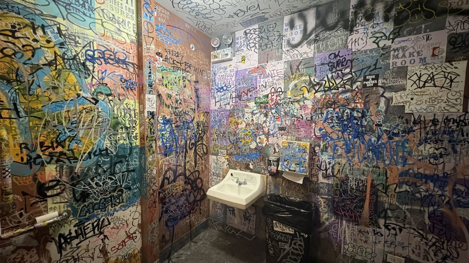 The bathroom is messy. Graffiti is scrawled on the walls. The various messages are written with blue, pink, yellow, green, black and red paint. There is an open trash can with a black liner that reflects the harsh lighting of the small restroom.